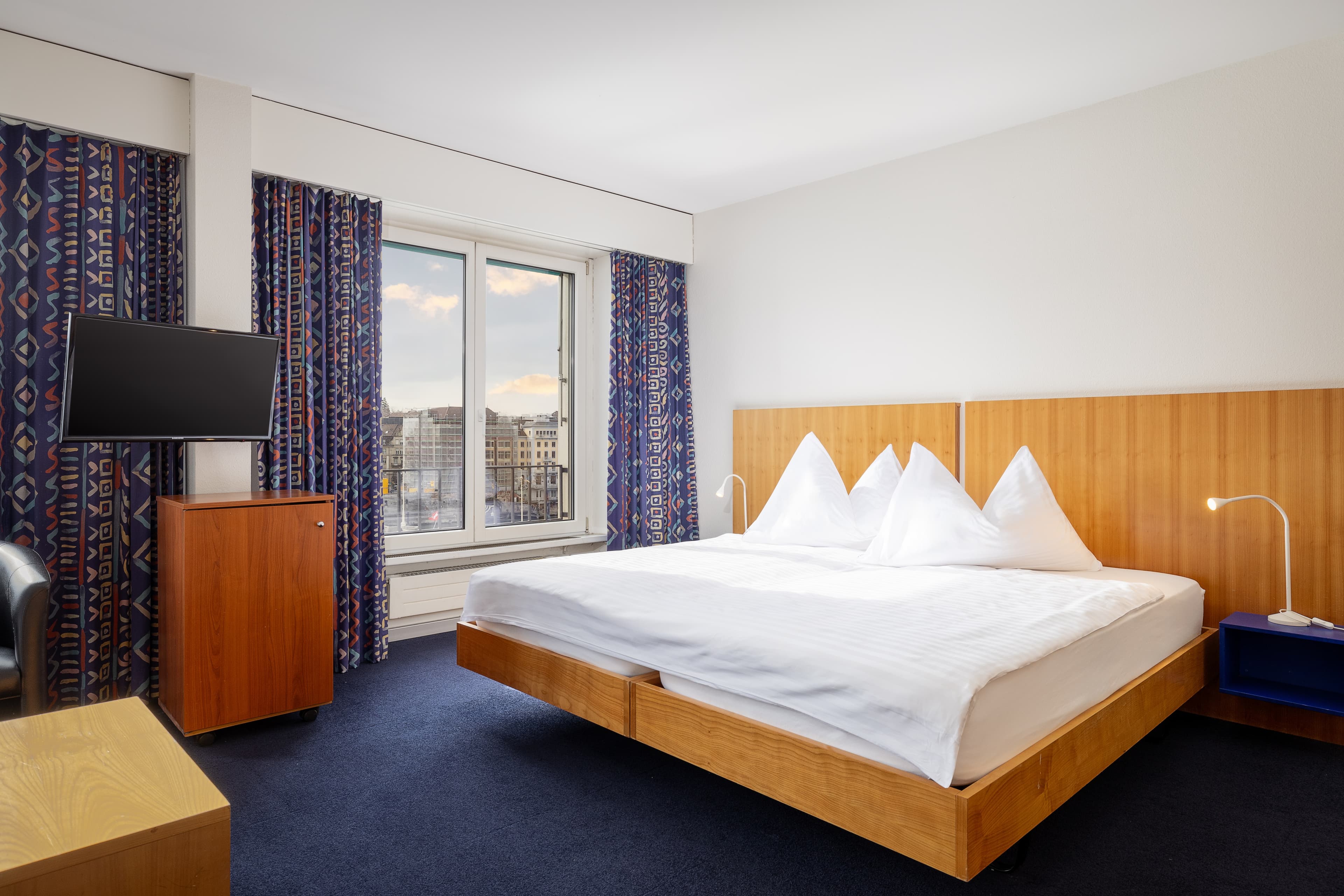 Junior Superior Suite at Hotel Merian in Basel with a modern double bed, large window with city view, flat screen TV, and stylish curtains.