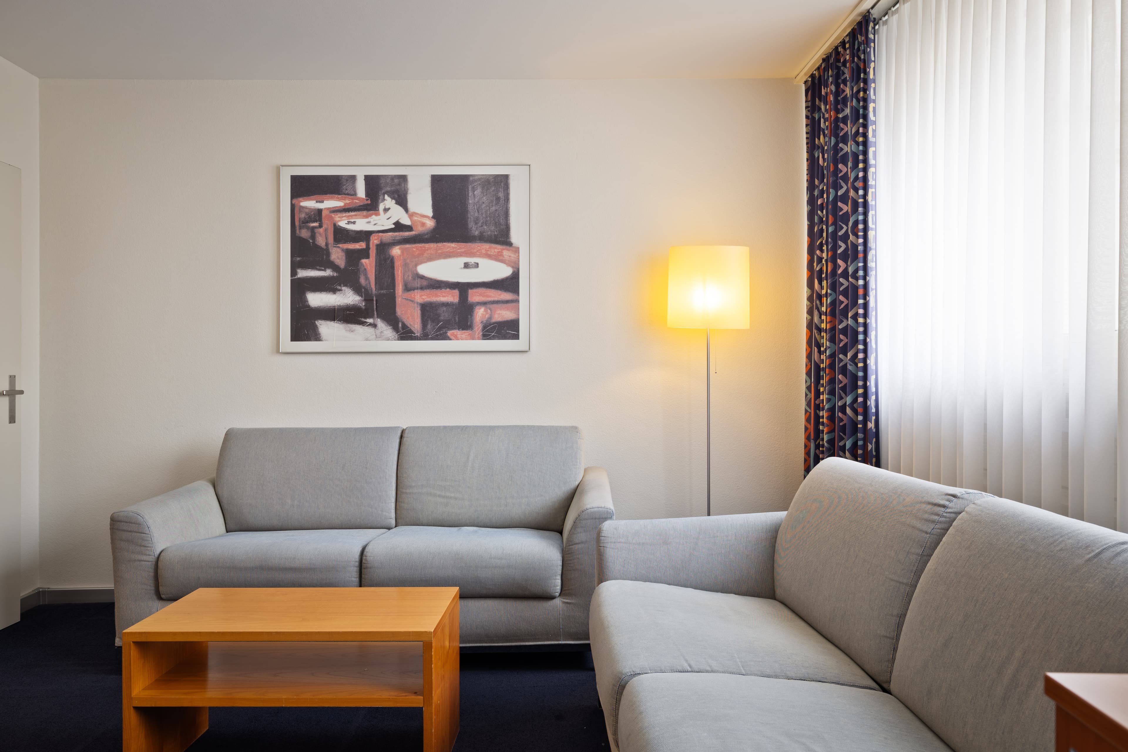 A living room of a Junior Suite Superior in the Hotel Merian Basel with two gray sofas, a wooden table, a floor lamp, and an art picture on the wall